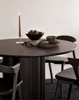 A modern round dark wood dining table with Ethnicraft Bok Dining Chairs in brown oak leather, set with stacked dark plates, a bowl, cup, brown napkin, and glass candle holder holding a lit brown taper candle. A fruit bowl sits at the center.