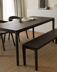 A minimalist dining area features a dark solid oak table, bench, and the Ethnicraft Bok Dining Chair in brown oak with leather on a woven rug. An open book and cup rest on the table. Neutral walls, curtains, and framed art create a calm, modern vibe.