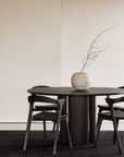 A minimalist dining area with a round dark wood table, three Ethnicraft Bok Dining Chairs in brown oak with leather, and a striped ceramic vase with bare branches, set on a black rug against a neutral wall.