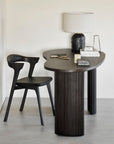 A modern desk with fluted legs is paired with the Ethnicraft Bok Dining Chair in brown oak and leather. On the desk are a textured-base lamp, stacked books, a metal sculpture, and decor—all set in a neutral, contemporary space.
