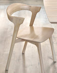 The Ethnicraft Bok Dining Chair - Oak, featuring a curved backrest and minimalist solid oak design, sits on a light wood floor beside a matching light wooden table.