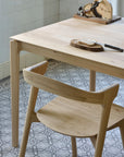 An Ethnicraft Bok Dining Chair - Oak pairs with a matching table on patterned tile flooring, topped with a vintage slicer, bread on a solid oak board, and a black-handled knife. White tiled walls complete the look.