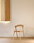 An Ethnicraft Bok Dining Chair - Oak rests on a light wood floor near a window with a partially open wooden shutter and a plain beige wall, as sunlight gently brightens the tranquil space.
