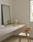 A minimalist workspace with a wooden floating desk, an Ethnicraft Bok Dining Chair - Oak, a sculptural lamp, standing mirror, cup and saucer, book, and pencil by the window in natural light.