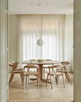 Minimalist dining room with a round solid oak table, six Ethnicraft Bok Dining Chair - Oak, a white pendant light, sheer curtains, and a bowl of green apples. The space has a bright neutral palette and chevron wood flooring.