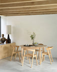 Minimalist dining area featuring a round solid oak table with four Ethnicraft Bok Dining Chair - Oak. A potted plant and tray rest on the table. A sideboard with vases and a lamp sits against a white wall beneath exposed wooden beams.