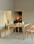 A minimalist dining room features a solid oak table, Bok Dining Chair - Oak by Ethnicraft, a matching bench, beige cushions, and a lit fireplace. Firewood is stacked below as soft natural light casts shadows on the pale wall.