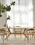 A modern dining room with a light Ethnicraft solid oak table, four Ethnicraft Bok Dining Chairs - Oak, a tall leafy plant, sheer white curtains, and sunlight streaming through large windows onto a pale rug and parquet floor.