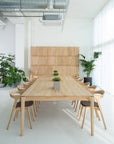 A modern, minimalist meeting room features a long solid oak table surrounded by eight Ethnicraft Bok Dining Chairs in oak with dark brown fabric, lush potted plants, a large window with sheer white curtains, and exposed ductwork on the ceiling.