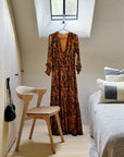 A long, orange floral dress hangs from a skylight window in a minimalist bedroom beside an Ethnicraft Bok Dining Chair - Oak with Leather, a bed with neutral pillows, and a small black handbag on the wall.