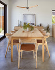 Bright dining room with a solid oak table and six Ethnicraft Bok Dining Chairs in oak with leather, a floral centerpiece, minimalist decor, large window on the left, and framed art against the back wall.