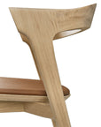Close-up of the Ethnicraft Bok Dining Chair - Oak with Leather, showing its curved backrest, light brown Nappa leather seat, clean lines, and smooth solid oak grain against a white background.