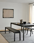 A minimalist dining room showcases the Bok Dining Table - Black by Ethnicraft with matching benches and chairs on a gray rug. Black dishes and glassware decorate the space, while light curtains frame the window and beige art hangs above.