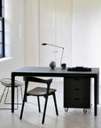 Minimalist office setup featuring the Ethnicraft Bok Dining Table - Black, paired with modern chairs, a rolling drawer unit, desk lamp, marble sphere, and window—all set in a bright, neutral space.