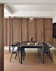 A modern dining room with the Bok Dining Table in brown oak by Ethnicraft, six matching black chairs, wooden walls, a ceramic vase, books and mugs on the table, and minimalist pendant lights above.