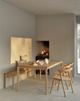 A minimalist dining room centers on the Ethnicraft Bok Dining Table - Oak, surrounded by four chairs and a cushioned bench. Sunlight enhances the warm European oak tones, while neatly stacked firewood rests below a lit fireplace.