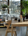 A sunlit modern dining area features Ethnicraft Bok Dining Table in oak, wooden chairs, and a bed by tall green plants near large windows with city views. Books from the Bok Collection and a potted plant sit on a black metal shelf nearby.