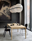 Modern dining area with the Ethnicraft Bok Dining Table in oak, two black chairs, three round pendant lights, and a large vintage-style wall portrait beside floor-to-ceiling windows.