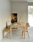 A minimalist dining area showcases the Ethnicraft Bok Dining Table in oak, paired with four matching chairs and a bench. A vase with bare branches decorates the table, while a modern fireplace adds warmth near a window overlooking snowy scenery.