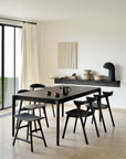 Minimalist dining room featuring an Ethnicraft Bok Extendable Dining Table in black with six matching chairs, neutral decor, ceramic bowls on a shelf, light tiled floor, white walls, and natural light from a large window with sheer curtains.