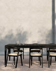 Minimalist outdoor dining area with an Ethnicraft table and six Bok Outdoor Dining Chairs in Black Teak, each with light cushions, arranged on a pebble ground against a textured light wall. A glass carafe and glass decorate the table.