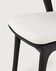 Close-up of the Ethnicraft Bok Outdoor Dining Chair, crafted with FSC-certified black teak legs and a textured off-white upholstered seat, shown against a simple light background.