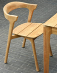 The Bok Outdoor Dining Chair by Ethnicraft features FSC-certified natural teak, a curved backrest, visible three-leg design, and smooth finish. Shown with a matching wooden table on gray tile as part of Ethnicrafts outdoor collection.