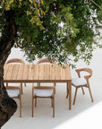 A Bok Outdoor Dining Table with six matching Ethnicraft Bok Outdoor Dining Chairs in natural teak, each with white cushions, sits on a white patio under a tree’s shade—crafted from FSC-certified teak.