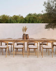 An Ethnicraft outdoor set featuring a long FSC-certified teak dining table and ten Bok Outdoor Dining Chairs in natural teak, with two vases of dried grasses, placed on a light patio against a white wall and greenery backdrop.