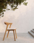 The Ethnicraft Bok Outdoor Dining Chair, crafted from FSC certified natural teak and featuring a light cushion, sits on a concrete patio beneath leafy branches, against a textured off-white wall.