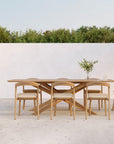 An Ethnicraft teak dining table with five Bok Outdoor Dining Chairs in Natural Teak sits on a patio by a white wall, decorated with a vase of flowers. Bushes and blue sky form the backdrop. All pieces are FSC certified from the Ethnicraft outdoor range.