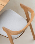 A top view of the Ethnicraft Bok Outdoor Dining Chair in natural teak with a curved backrest and light gray upholstered seat, placed next to a matching table from the Ethnicraft collection on a light-colored floor.