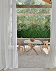 A modern outdoor dining area features a wooden table, four Ethnicraft Bok Outdoor Dining Chairs in natural teak, a bowl centerpiece, gravel flooring, sheer white curtains, and lush greenery in the background.