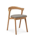 The Bok Outdoor Dining Chair by Ethnicraft features a minimalist design with angled legs, a curved backrest, FSC certified natural teak construction, and a light gray cushioned seat—perfect for stylish outdoor settings.