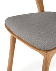 A close-up of the Ethnicraft Bok Outdoor Dining Chair in natural teak, featuring a light brown FSC-certified teak frame and a textured gray padded seat, showing part of the seat and two legs.