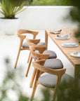 Three modern wooden chairs with gray cushions are arranged beside the Ethnicraft Bok Outdoor Dining Table in natural teak, set with white plates, glasses, and folded napkins on a sunny patio with greenery in the background.
