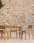 The Ethnicraft Bok Outdoor Dining Table in natural teak, paired with three matching chairs, is set on a stone patio with a bowl of fruit and plates, against a rustic stone wall backdrop.