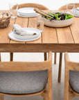 The Ethnicraft Bok Outdoor Dining Table in natural teak is elegantly set with white plates, gray napkins, clear glasses, a bread basket, and salad in a large bowl with wooden utensils. Four cushioned wooden chairs complete this FSC certified ensemble.