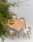 The Ethnicraft Bok Outdoor Dining Table in natural teak is paired with three curved wooden chairs on a light floor. A wooden bowl of yellow lemons and a pomegranate sits atop, while green branches with yellow fruit hang nearby.