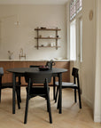 Minimalistic dining room featuring the Ethnicraft Bok Round Extendable Dining Table in black, four black chairs, light wood floors, a wooden kitchen cabinet, open shelves with simple dishes, and large windows filling the space with natural light.