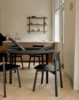 A minimalist kitchen with light wood floors, black chairs, wall shelves with ceramics, and a brass faucet features an Ethnicraft Bok Round Extendable Dining Table in Black. A person adjusts an item on the table as natural light fills the space.