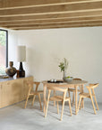 Minimalist dining area with an Ethnicraft Bok Round Extendable Dining Table in oak, four matching chairs, a sideboard topped with vases and a lamp, plus a bowl of branches—all set against white walls and exposed wooden beams.