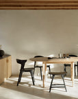 Minimalist dining area with an Ethnicraft Bok Round Extendable Dining Table in oak, four black chairs, and sunlight. A sideboard with neutral-toned decor and exposed wooden ceiling beams complete the warm, modern look.