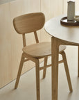 A light wooden chair with a curved backrest stands by Ethnicraft’s Bok Round Extendable Dining Table in oak, which features a tray with a pitcher and bowl. The scene reflects minimalist, natural style.