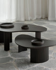 Two Boomerang Coffee Tables by Ethnicraft, crafted from varnished mahogany with curved edges and ribbed cylindrical legs, stand on a stone-tiled floor. Each displays a black dish, framed by sheer white curtains in the background.