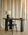 The Ethnicraft Boomerang Desk with ribbed legs in black sits before beige curtains, paired with a black chair. On the desk are a black lamp with white shade, books, a pen, mug, and decorative accents.