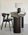 A modern workspace with Ethnicrafts Boomerang Desk, fluted legs, a black sculptural chair, ceramic lamp, and books—classic European refinement against a light wall.