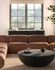 A modern living room with minimalist appeal features a brown leather sectional, two round black coffee tables, the Moes Insitu Side Table - Black, a large indoor tree, a sideboard, and tall windows that fill the space with natural light.
