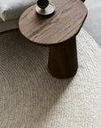 A small, round wooden side table with a dark finish holds a cup and saucer. It rests on the Armadillo&Co Braid Rug - Chalk, a textured, light-colored handmade wool-blend rug beside a light gray upholstered sofa.