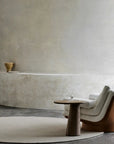 A minimalist room with a textured beige wall, modern cream armchair, round wooden side table with water, and the Braid Rug - Chalk by Armadillo&Co. A small sculptural bowl sits on a ledge in the background.